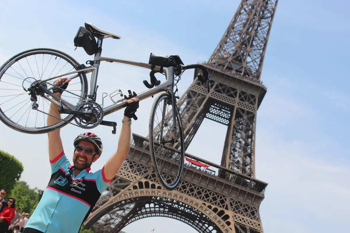 A Cambridge Community Church (C3) charity rider lifting his bike above his head under the Eiffel Tower