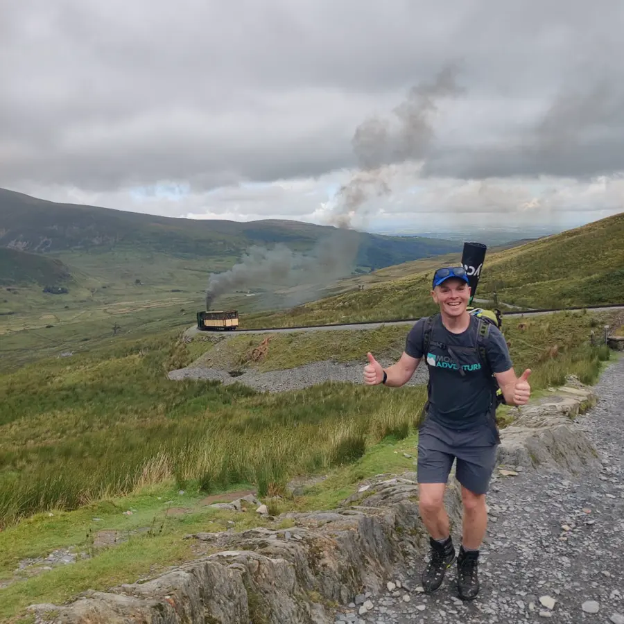 Tom giving a thumbs up on the path up Yr Wyddfa, with the mountain railway behind