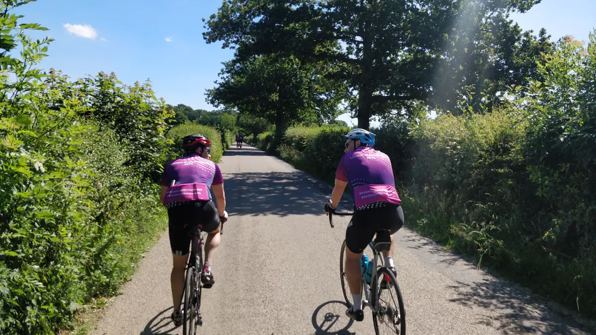 Two riders in team jerseys cycling along a sunny tree-lined lane